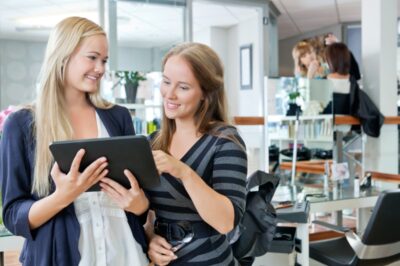 Two young, blonde woman talking over a tablet computer in a hair salon