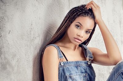 Beautiful, young black woman with natural hair braids and overalls sitting against a grey wall