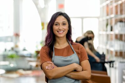 Young, smiling woman with dark hair highlighted purple standing in foreground of salon
